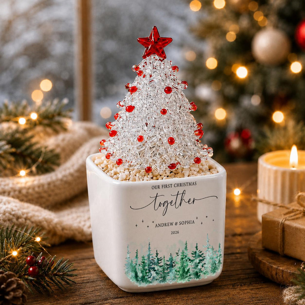 May include: A clear glass Christmas tree ornament with red accents and a red star topper, set in a white square ceramic planter. The planter features the text "OUR FIRST CHRISTMAS together ANDREW & SOPHIA 2026" and a green watercolor pine tree design.