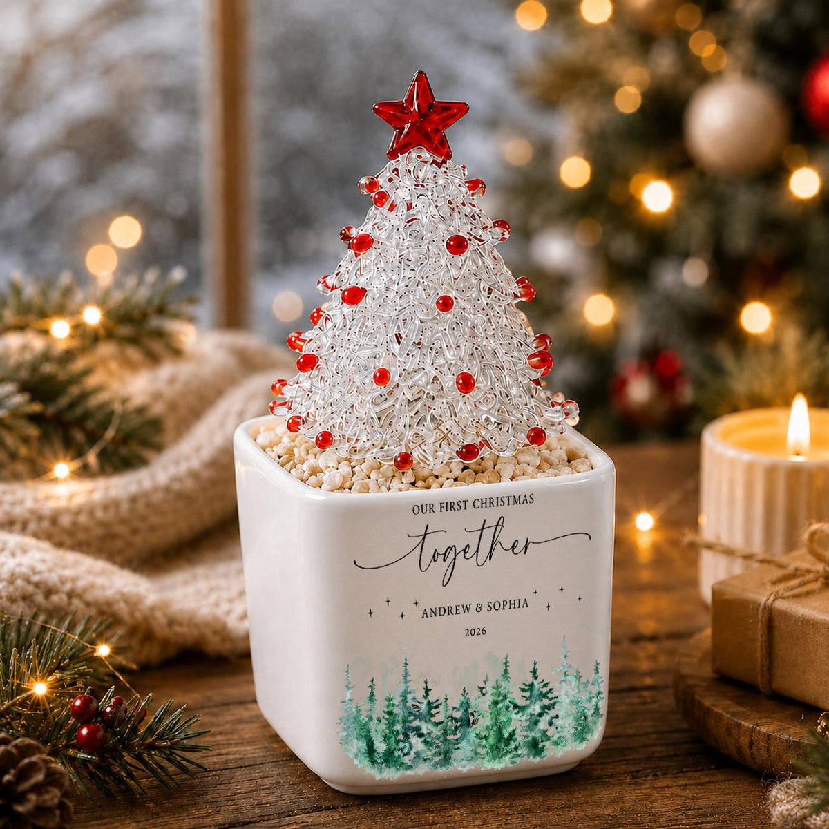 May include: A clear glass Christmas tree ornament with red accents and a red star topper, set in a white square ceramic planter. The planter features the text "OUR FIRST CHRISTMAS together ANDREW & SOPHIA 2026" and a green watercolor pine tree design.