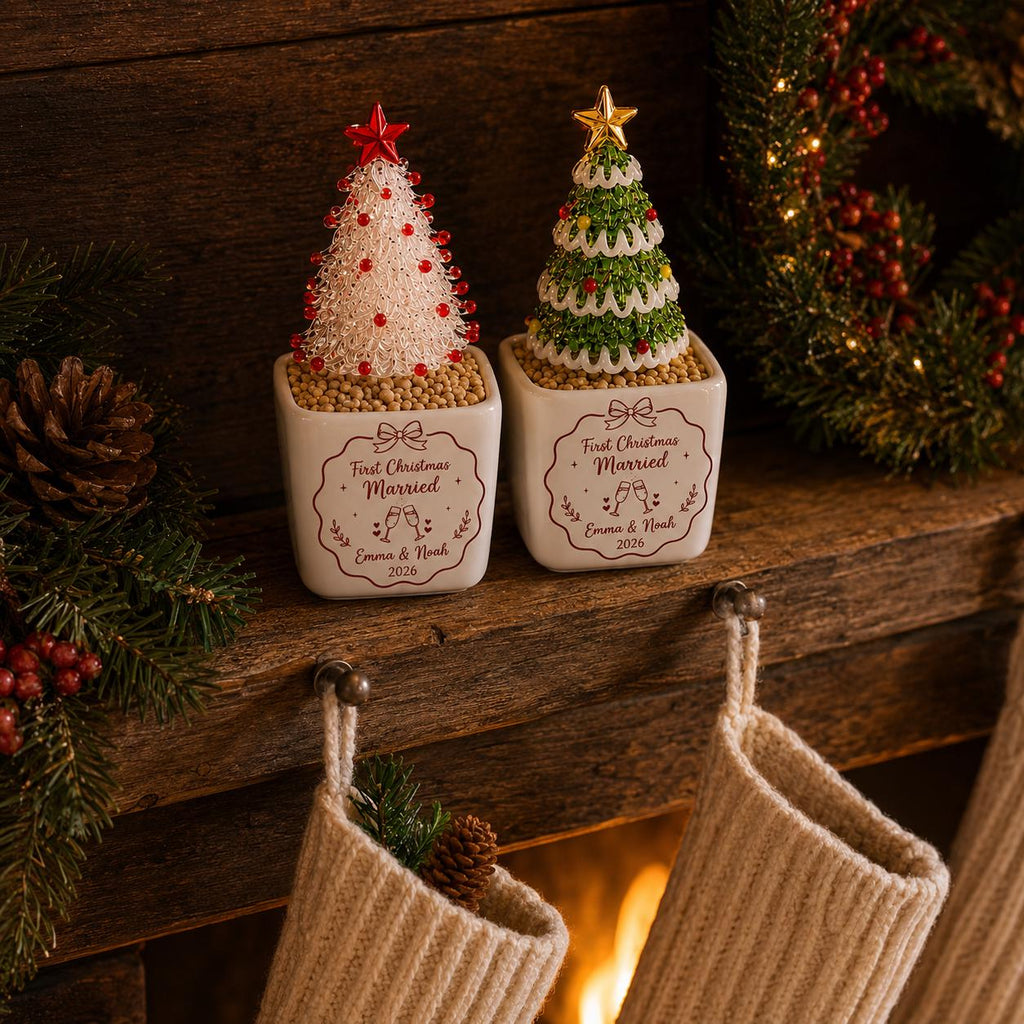 May include: Two square white ceramic planters containing miniature Christmas trees. One tree is white with red beads and a red star, the other is green with white trim and a gold star. Each planter features the text First Christmas Married, Emma & Noah, 2026.