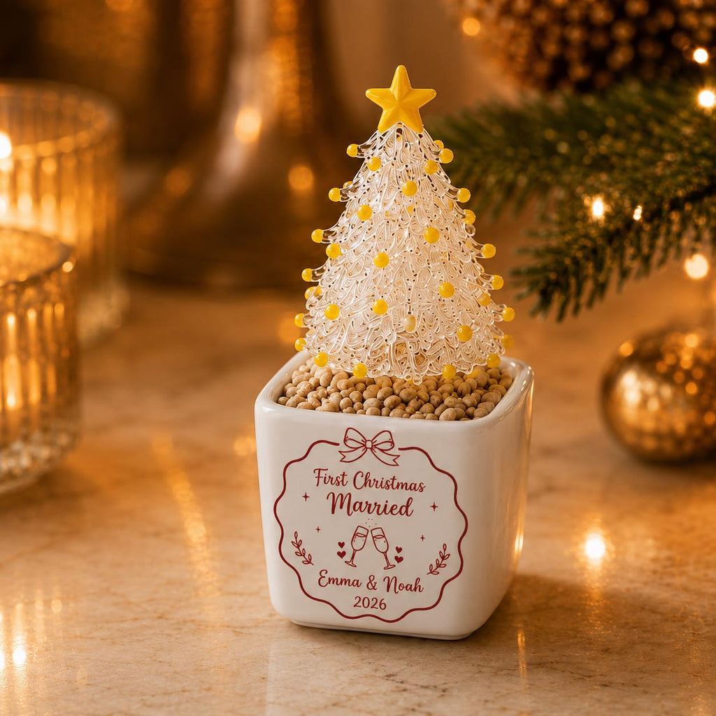 May include: A clear glass Christmas tree ornament with yellow accents and a yellow star topper, set in a white square ceramic planter filled with small tan pebbles. The planter features red text reading First Christmas Married, Emma & Noah, 2026.