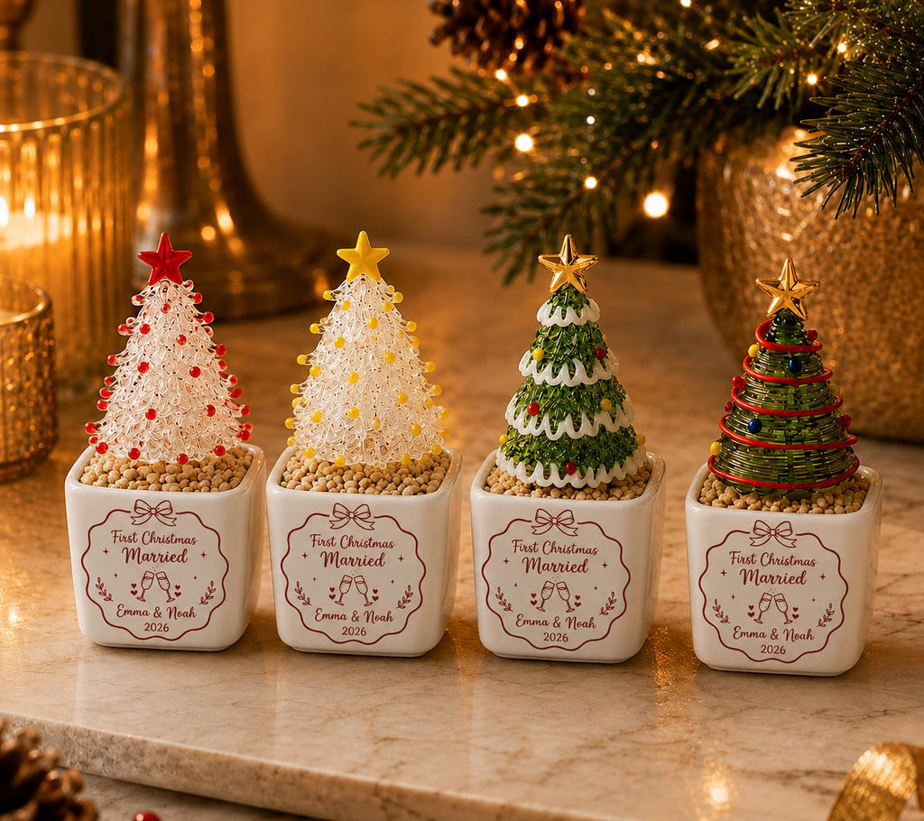 May include: Four miniature decorative Christmas trees in square white ceramic pots. Each pot features the text First Christmas Married, Emma & Noah, and 2026. The trees vary in design, featuring red, yellow, green, and dark green colors with stars.