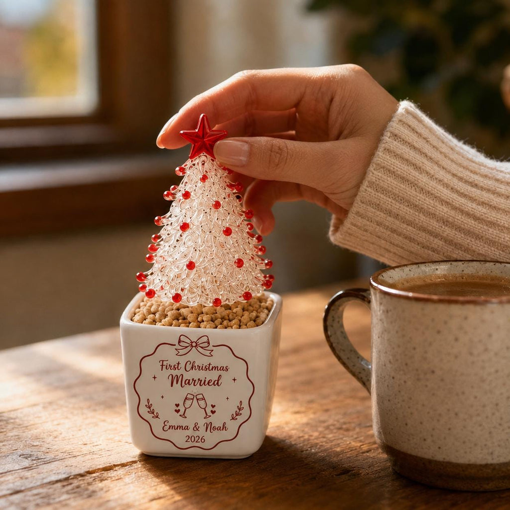 May include: A white square ceramic planter filled with small tan pebbles, topped with a clear glass Christmas tree ornament featuring red bead accents and a red star. The planter displays the text First Christmas Married, Emma & Noah 2026.