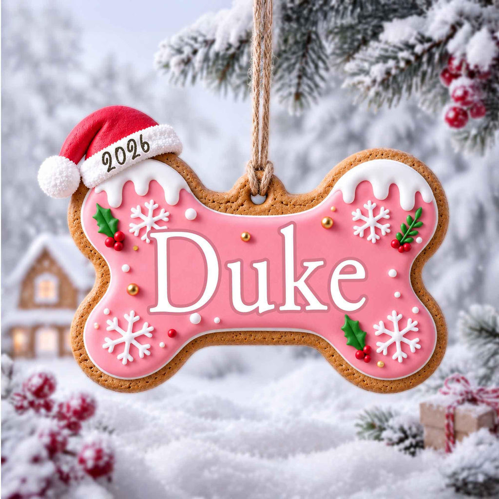 May include: A bone-shaped gingerbread ornament with pink icing, white snowflake patterns, and green holly accents. The name Duke is written in white letters. A red Santa hat with the year 2026 sits on the top left corner. It hangs from a jute twine.