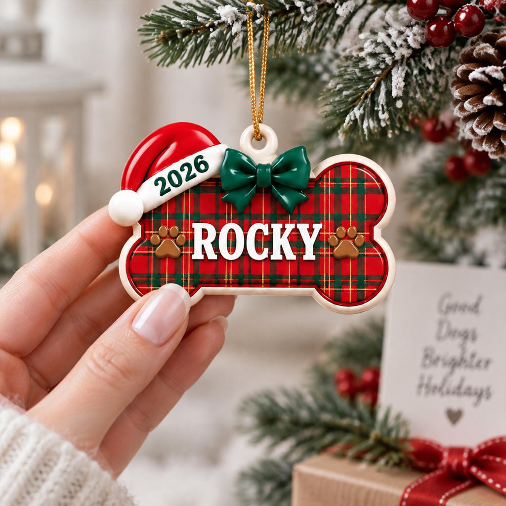 May include: A bone-shaped Christmas ornament featuring a red and green plaid pattern, a white Santa hat with the text 2026, a green bow, and the name ROCKY printed in white letters with two brown paw prints on a cream-colored border with a gold string.