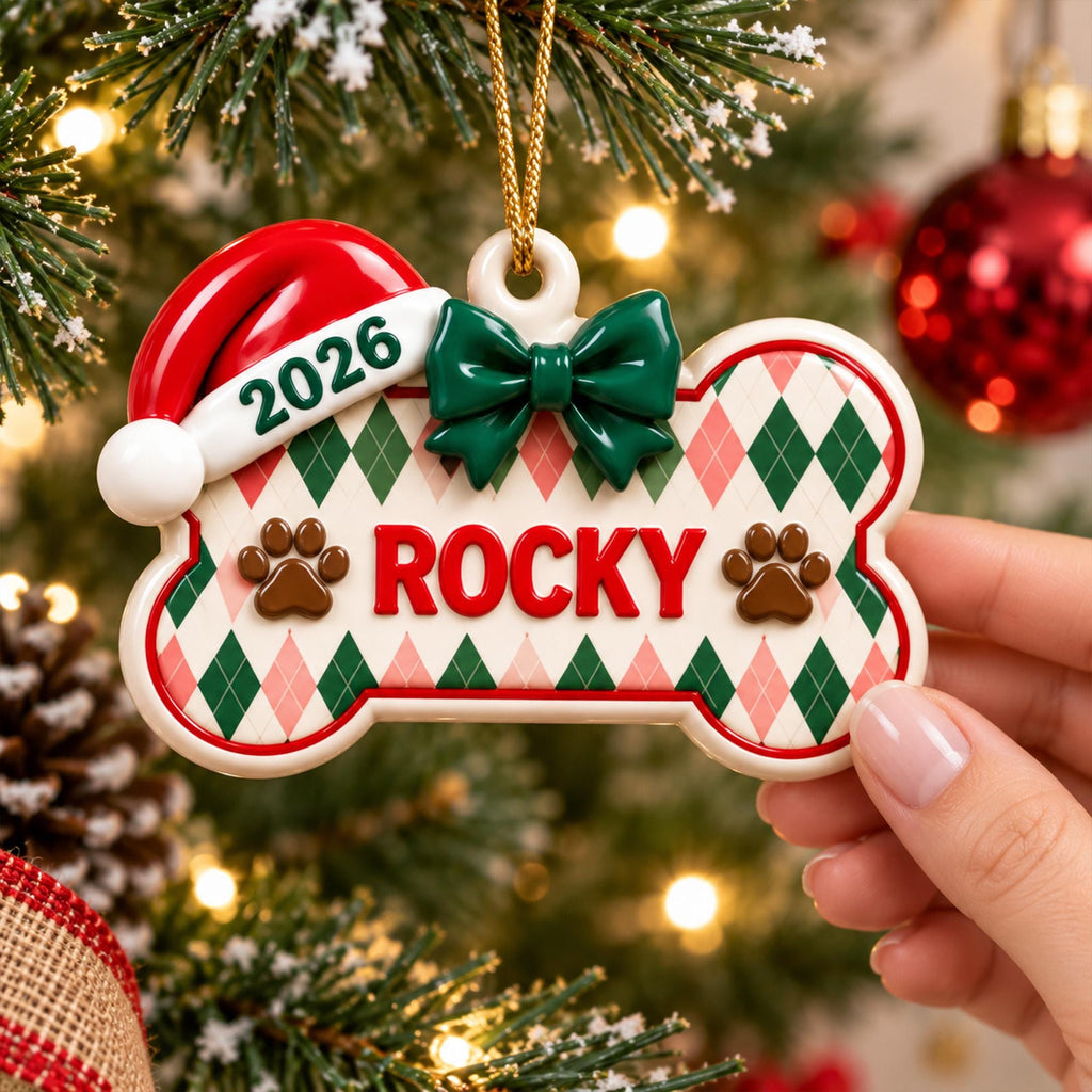 May include: A bone-shaped Christmas ornament featuring a cream background with a green and pink argyle pattern. The ornament displays the name ROCKY in red letters, two brown paw prints, a green bow, and a red Santa hat with the year 2026 printed.