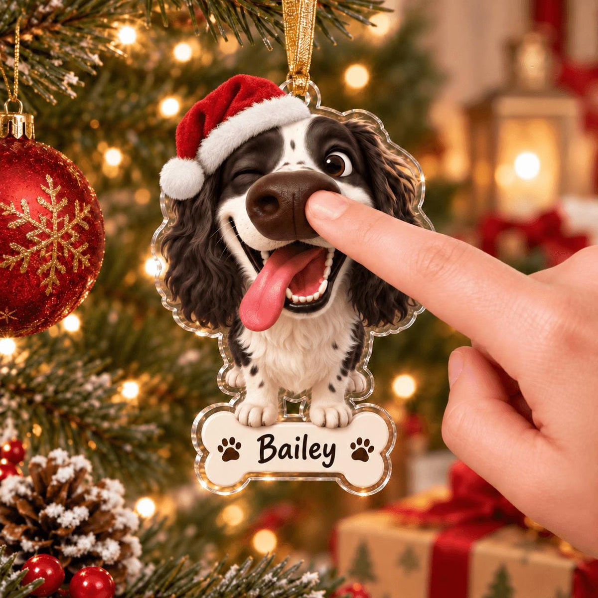 May include: A clear acrylic Christmas ornament shaped like a winking black and white spaniel dog wearing a red Santa hat with a white pom-pom. The dog has its tongue out and is attached to a bone-shaped name tag with the printed name Bailey.