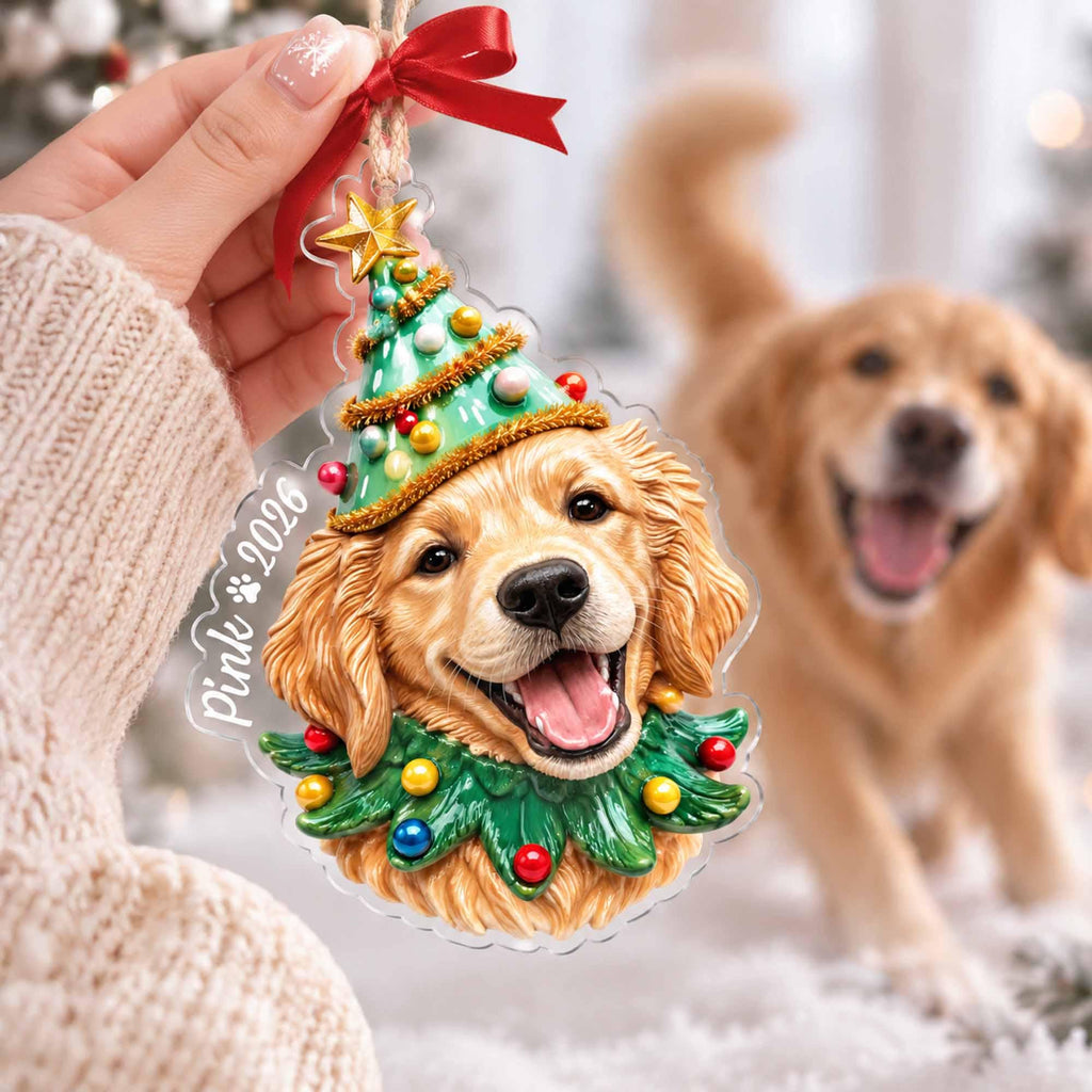 May include: A clear acrylic Christmas ornament featuring a golden retriever wearing a green cone-shaped hat with a gold star and a green collar, both decorated with colorful spheres. The text Pink 2026 is printed on the side with a red ribbon.