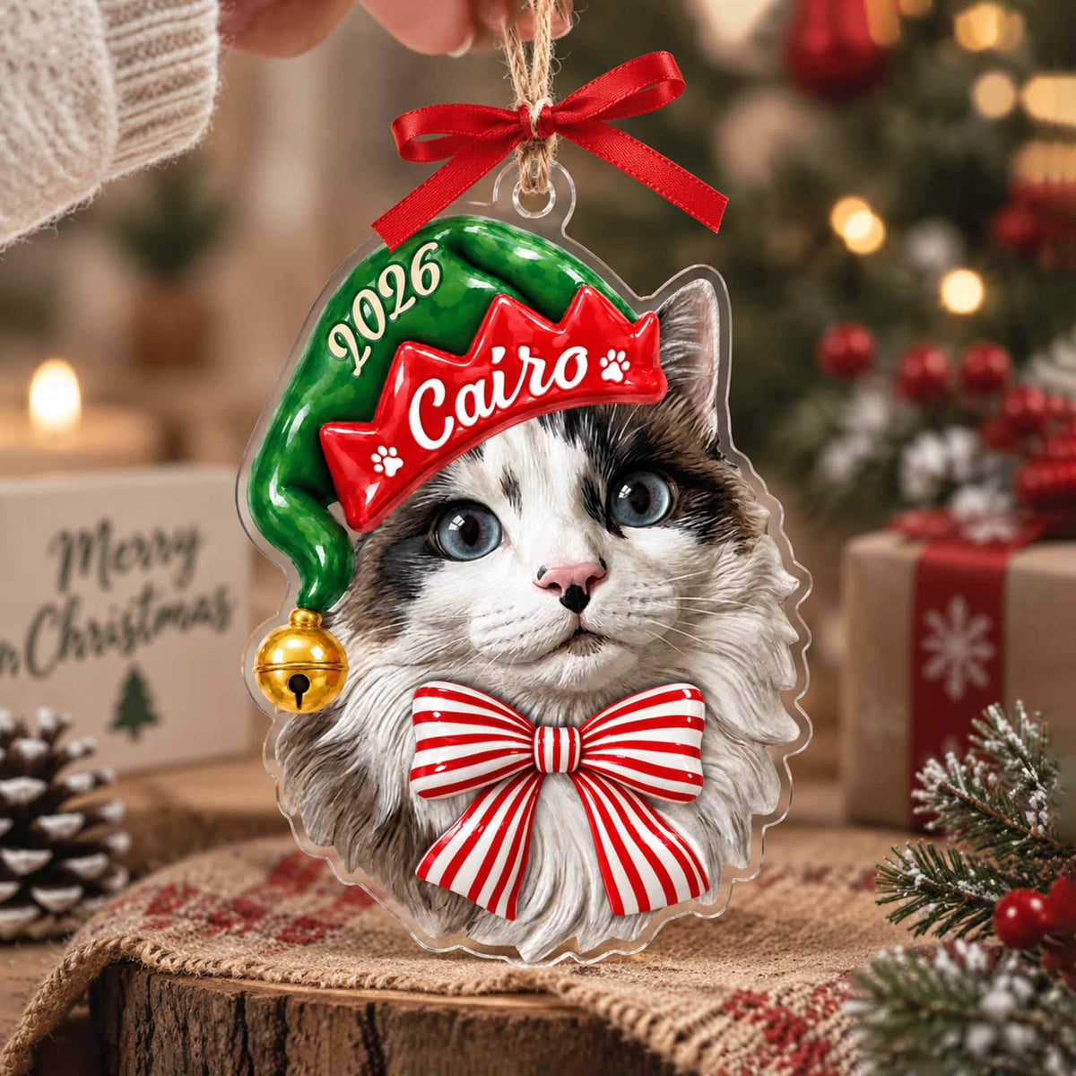 May include: A clear acrylic Christmas ornament featuring a cat wearing a green elf hat with the text 2026 and Cairo printed on it, accented by a small gold bell and a red ribbon bow at the top, and a large red and white striped bow at the bottom.