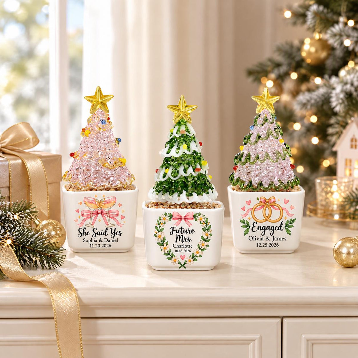 May include: Three small decorative Christmas trees in white square planters. The left tree is pink with "She Said Yes, Sophia & Daniel, 11.20.2026". The center is green with "Future Mrs., Charlotte, 10.18.2026". The right is green and pink with "Engaged, Olivia & James, 12.25.2026".