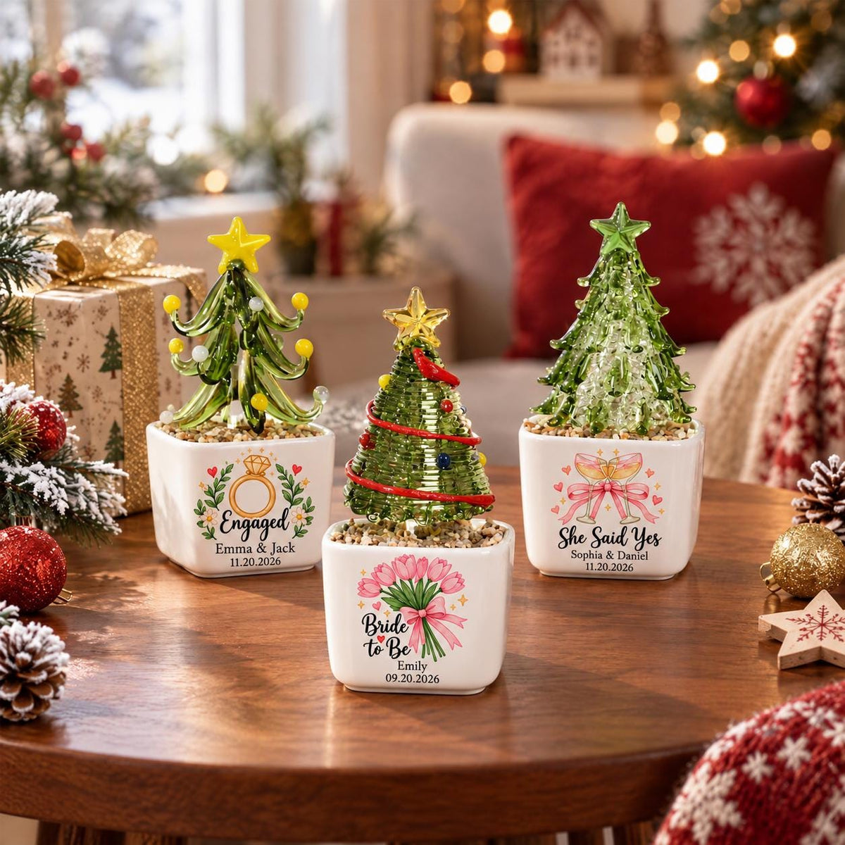 May include: Three glass Christmas tree ornaments in white square planters. The left tree has yellow accents and says Engaged Emma & Jack 11.20.2026. The center tree has red garland and says Bride to Be Emily 09.20.2026. The right tree says She Said Yes Sophia & Daniel 11.20.2026.