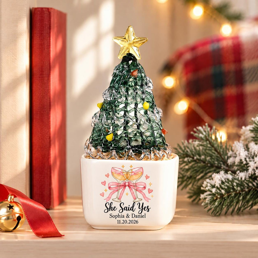 May include: A glass Christmas tree ornament with a gold star topper, sitting in a white square ceramic pot. The pot features a graphic of two champagne glasses with a pink bow, the text "She Said Yes", "Sophia & Daniel", and "11.20.2026".