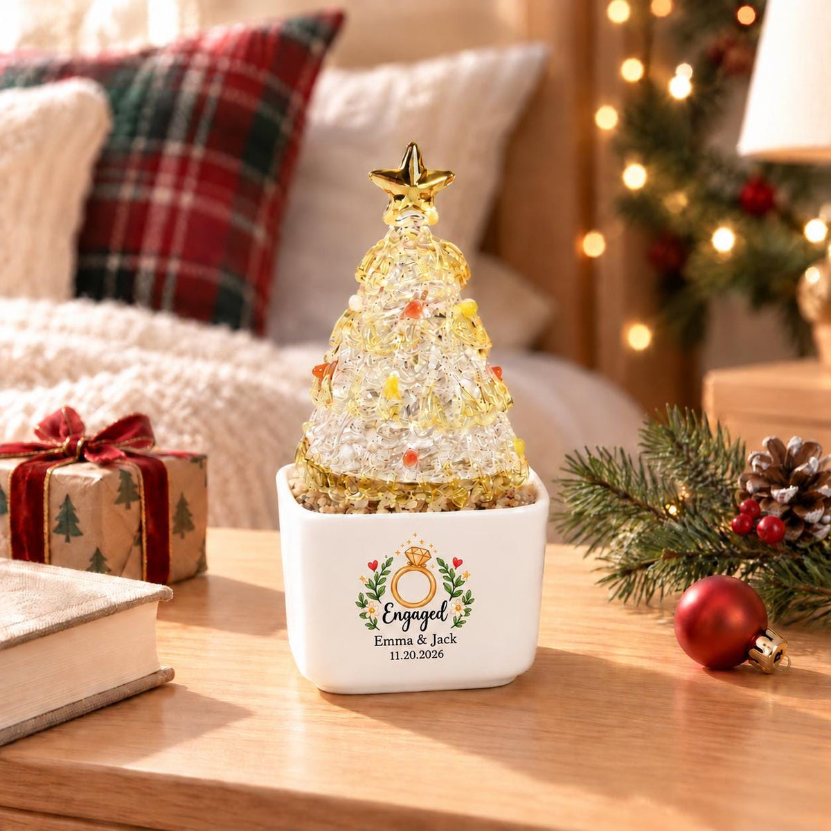 May include: A clear glass Christmas tree ornament with yellow and orange accents, topped with a gold star, sitting in a white square ceramic planter. The planter features a printed graphic of a ring, the text "Engaged", "Emma & Jack", and "11.20.2026".