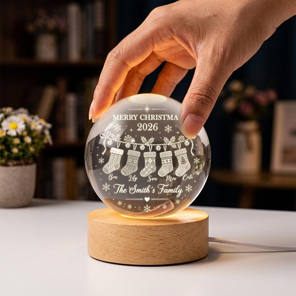 May include: A clear spherical glass ornament featuring an engraved design of five hanging Christmas stockings, the text MERRY CHRISTMA 2026, and the phrase The Smith's Family, resting on a round light-colored wooden base with an integrated light.