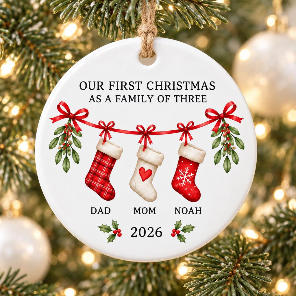 May include: A round white ceramic Christmas ornament featuring the text OUR FIRST CHRISTMAS AS A FAMILY OF THREE. It displays three hanging stockings with red ribbons and mistletoe, labeled DAD, MOM, and NOAH, with the year 2026 printed below.