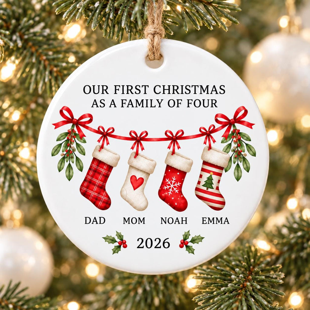 May include: A round white ceramic ornament hanging from a jute string. It features the text "OUR FIRST CHRISTMAS AS A FAMILY OF FOUR" above four hanging stockings with red bows and mistletoe. Below are the names "DAD", "MOM", "NOAH", "EMMA" and "2026".