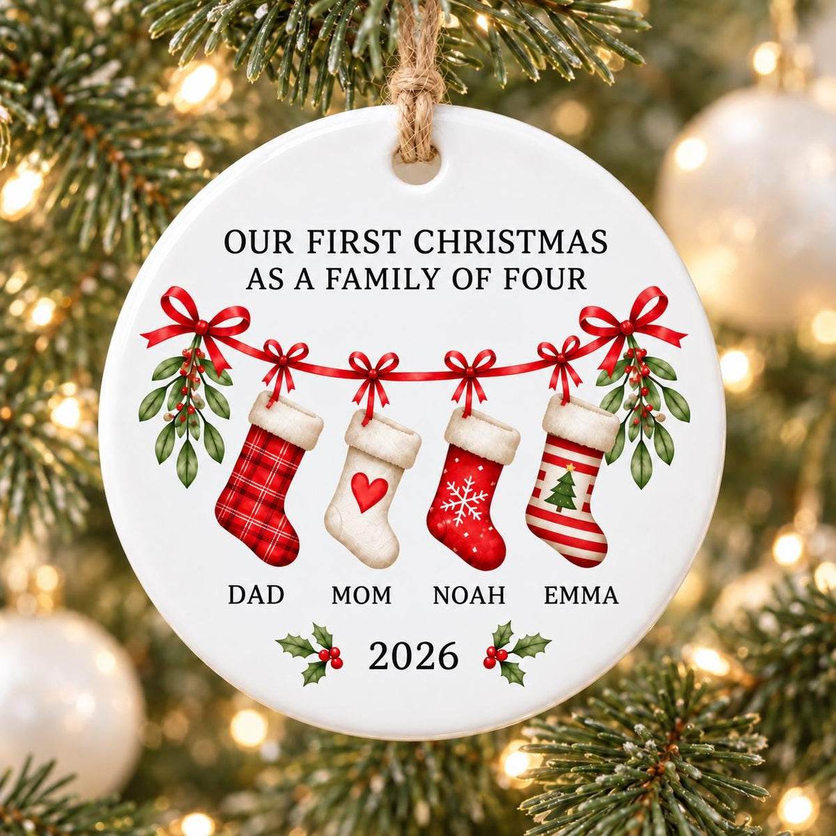 May include: A round white ceramic ornament hanging from a jute string. It features the text "OUR FIRST CHRISTMAS AS A FAMILY OF FOUR" above four hanging stockings with red bows and mistletoe. Below are the names "DAD", "MOM", "NOAH", "EMMA" and "2026".