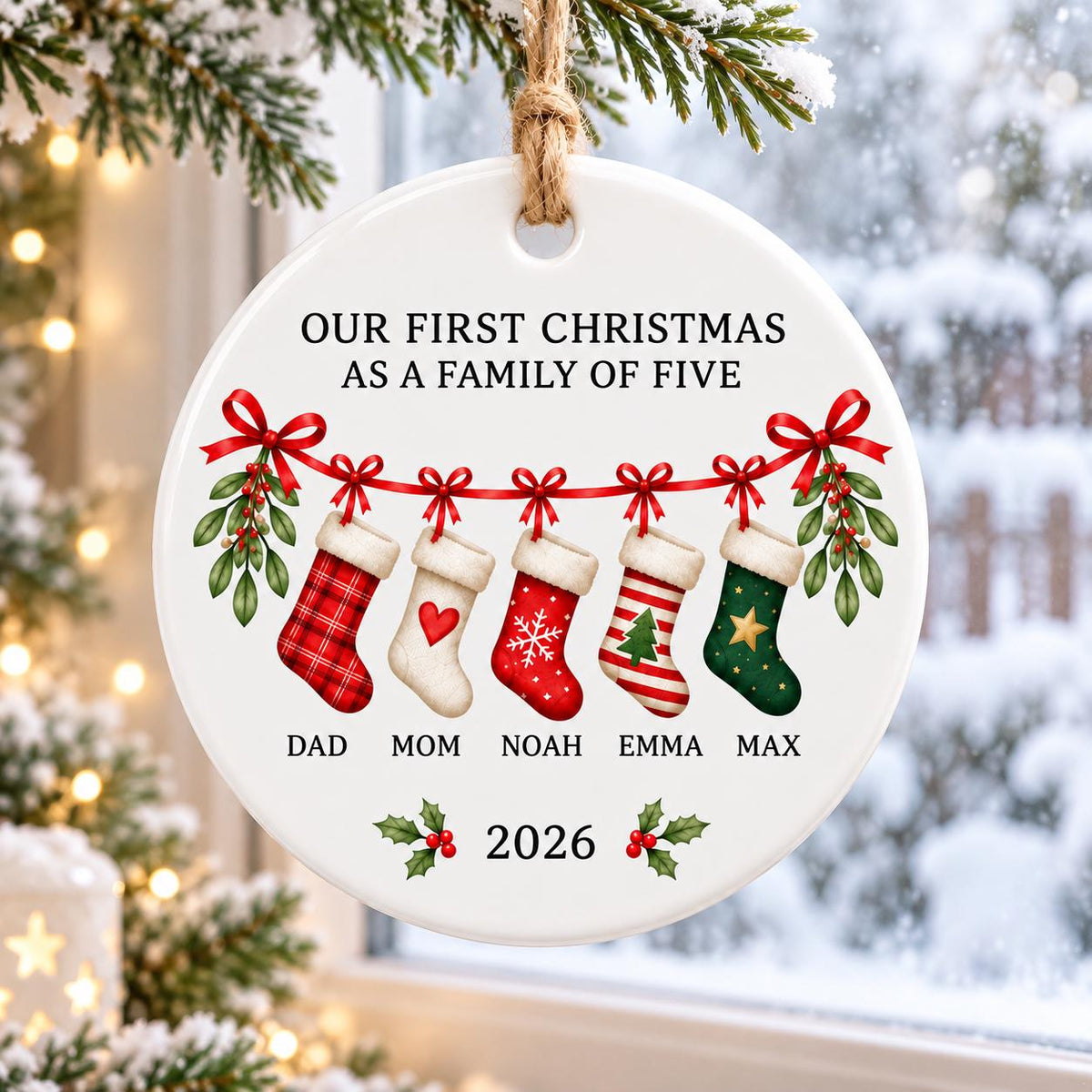 May include: A round white ceramic ornament hanging from a jute string. It features the text "OUR FIRST CHRISTMAS AS A FAMILY OF FIVE" above five hanging stockings with red bows and mistletoe. Names "DAD", "MOM", "NOAH", "EMMA", "MAX" and "2026" are printed.