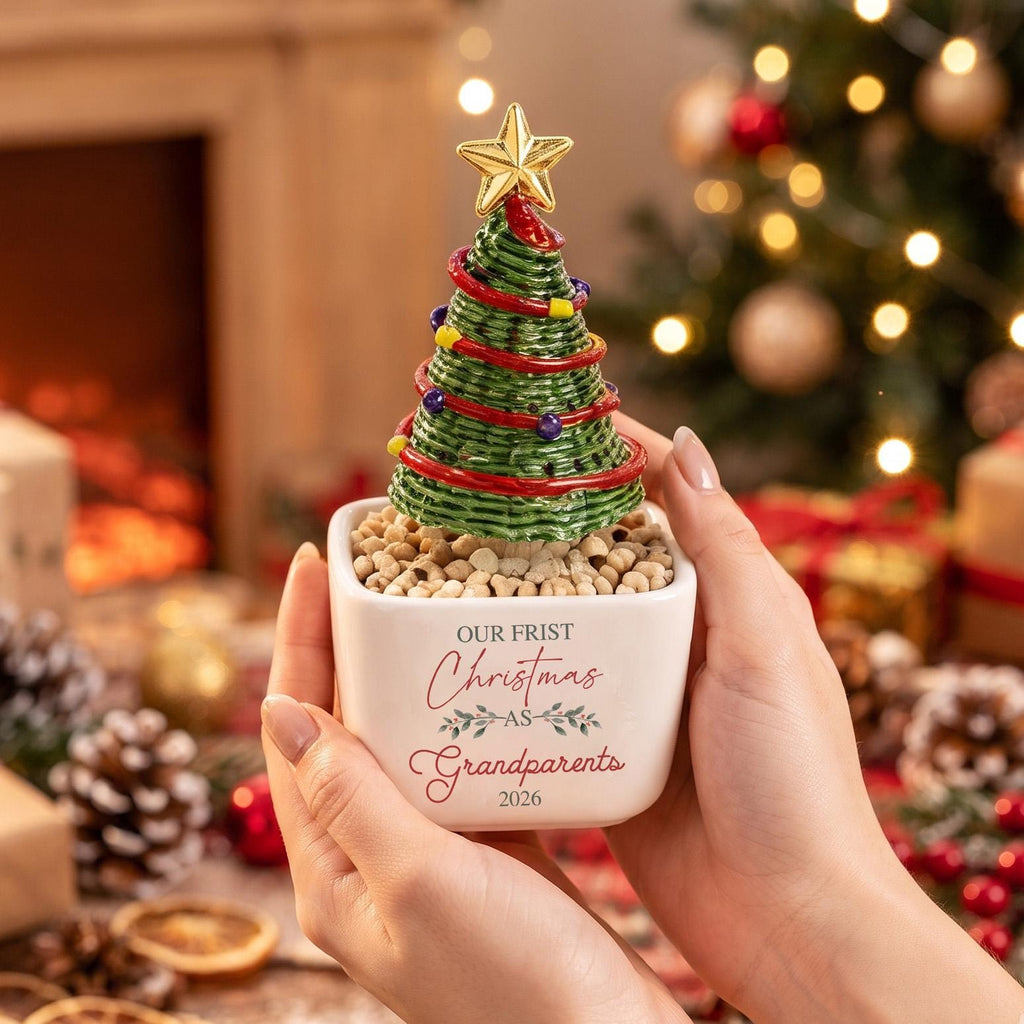 May include: A small, white, square ceramic planter filled with light-colored pebbles and a green, coiled-wire Christmas tree ornament. The tree features red wire garland, colorful beads, and a gold star topper. The planter reads: OUR FRIST Christmas AS Grandparents 2026.