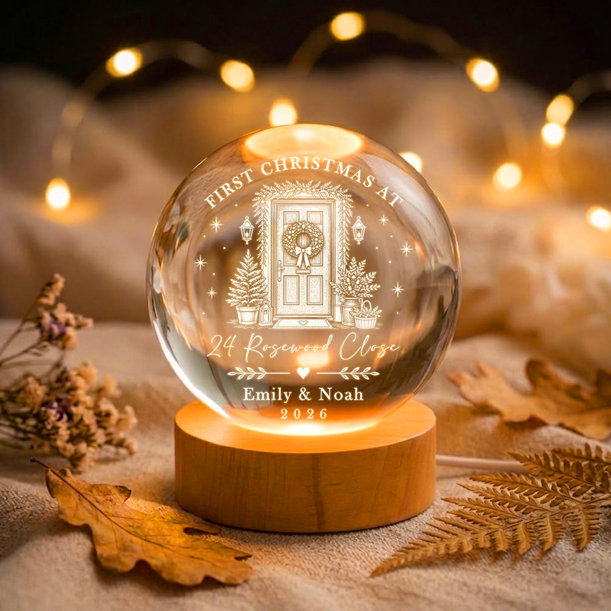 May include: A clear glass sphere ornament on a round wooden base, featuring an engraved illustration of a front door with a wreath, flanked by potted trees. Text reads: FIRST CHRISTMAS AT 24 Rosewood Close, Emily & Noah, 2026, with a small heart.