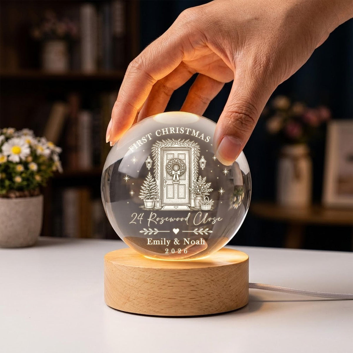 May include: A clear spherical glass ornament featuring an engraved illustration of a front door with a wreath, Christmas trees, and the text "FIRST CHRISTMAS," "24 Rosewood Close," "Emily & Noah," and "2026," mounted on a round wooden base.