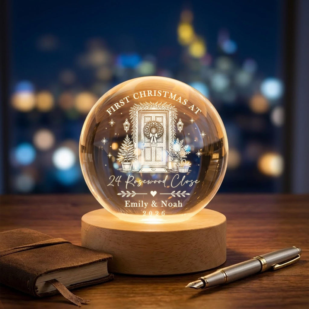 May include: A clear glass sphere ornament on a round wooden base, illuminated from within. The glass features an etched design of a front door with a wreath, stars, and the text: FIRST CHRISTMAS AT 24 Rosewood Close, Emily & Noah, 2026.