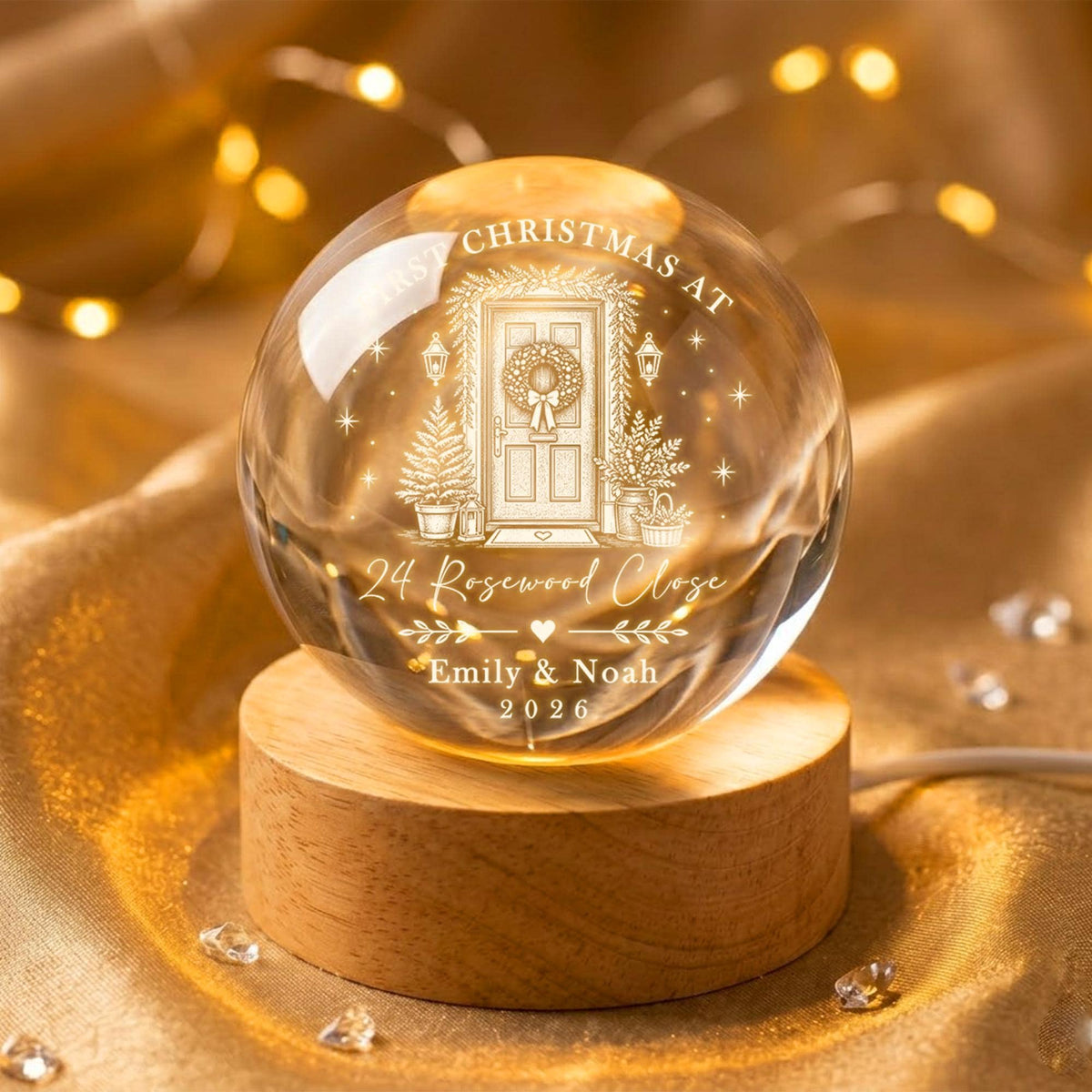 May include: A clear glass sphere resting on a cylindrical wooden base. The sphere features an etched design of a front door with a wreath, surrounded by stars and text reading FIRST CHRISTMAS AT 24 Rosewood Close EMILY & NOAH 2026.