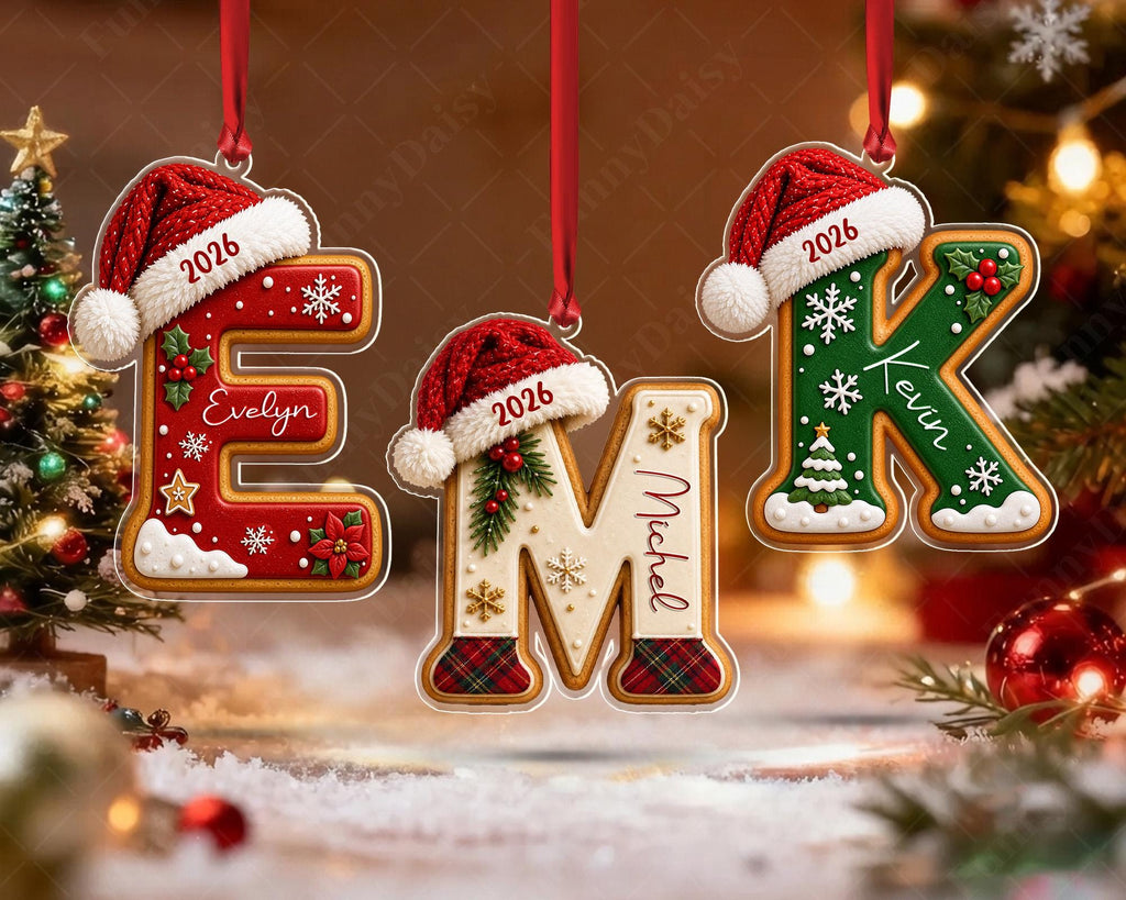 May include: Three hanging Christmas ornaments shaped like gingerbread letters E, M, and K. Each letter wears a red Santa hat with the year 2026 and a name: Evelyn, Michel, and Kevin. The letters feature festive decorations like snowflakes, holly, and plaid.