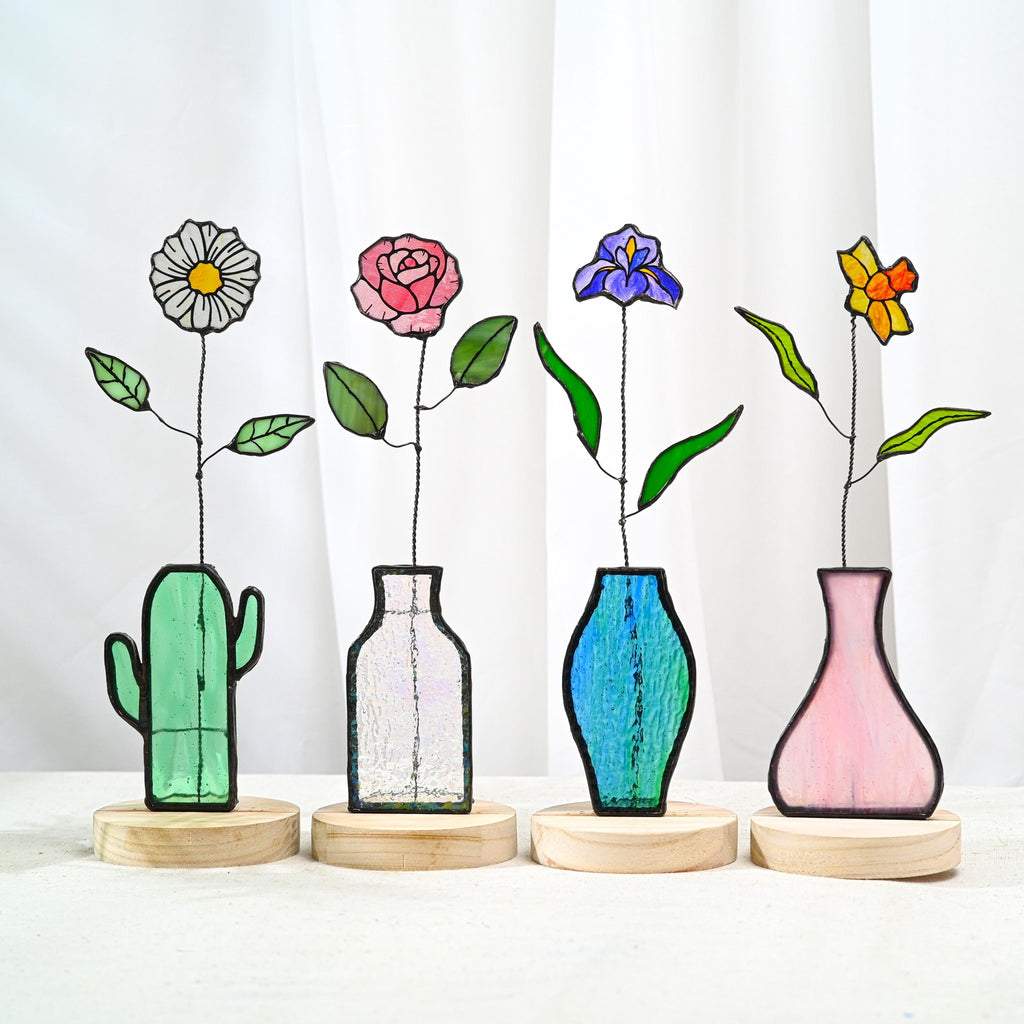 May include: Four stained glass flower sculptures on round wooden bases. The flowers include a white daisy, a pink rose, a purple iris, and a yellow daffodil. Each flower is held in a unique glass vase: a green cactus, clear, blue, and pink.
