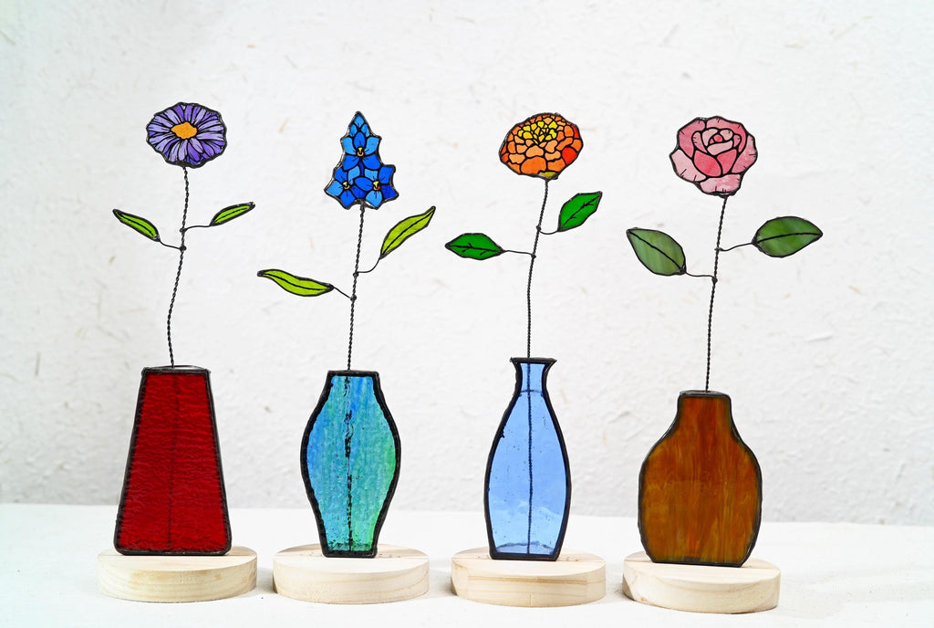 May include: Four stained glass flower sculptures on round wooden bases. Each features a unique flower: a purple daisy, a blue delphinium, an orange marigold, and a pink rose, each with wire stems and green glass leaves in distinct glass vases.