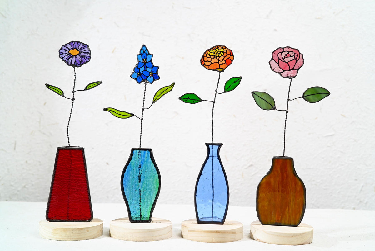 May include: Four stained glass flower sculptures on round wooden bases. Each features a unique flower: a purple daisy, a blue delphinium, an orange marigold, and a pink rose, each with wire stems and green glass leaves in distinct glass vases.