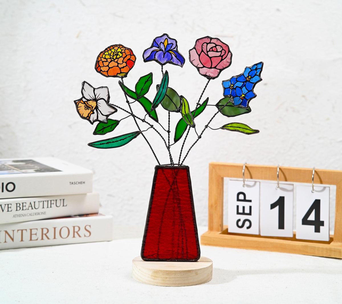 May include: A stained glass flower arrangement featuring a white daffodil, an orange marigold, a purple iris, a pink rose, and blue blossoms. The flowers have green leaves and wire stems, set in a red trapezoidal vase on a round wooden base.