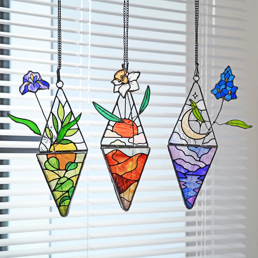 May include: Three diamond-shaped stained glass suncatchers hanging from black chains. The left features a purple iris and yellow foliage. The center displays a white daffodil over a red desert landscape. The right shows blue flowers above a crescent moon.