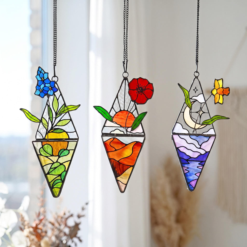 May include: Three diamond-shaped stained glass suncatchers hanging from black chains. The first features blue flowers and green leaves over a yellow sun. The second shows a red poppy above an orange landscape. The third depicts a crescent moon.