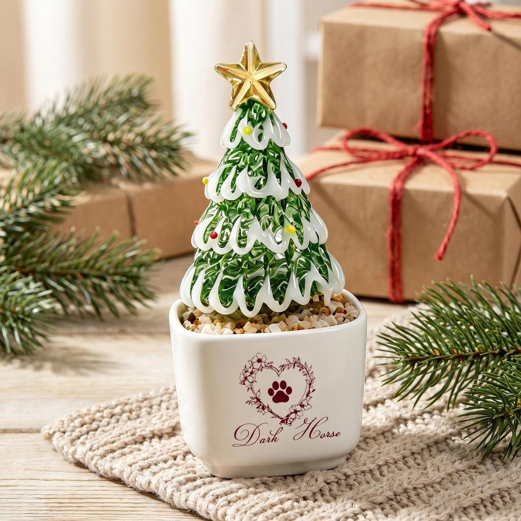 May include: A glass Christmas tree ornament with green and white swirls, topped with a gold star, sitting in a white square ceramic pot. The pot features a heart-shaped floral design with a paw print and the text Dark Horse printed in red.