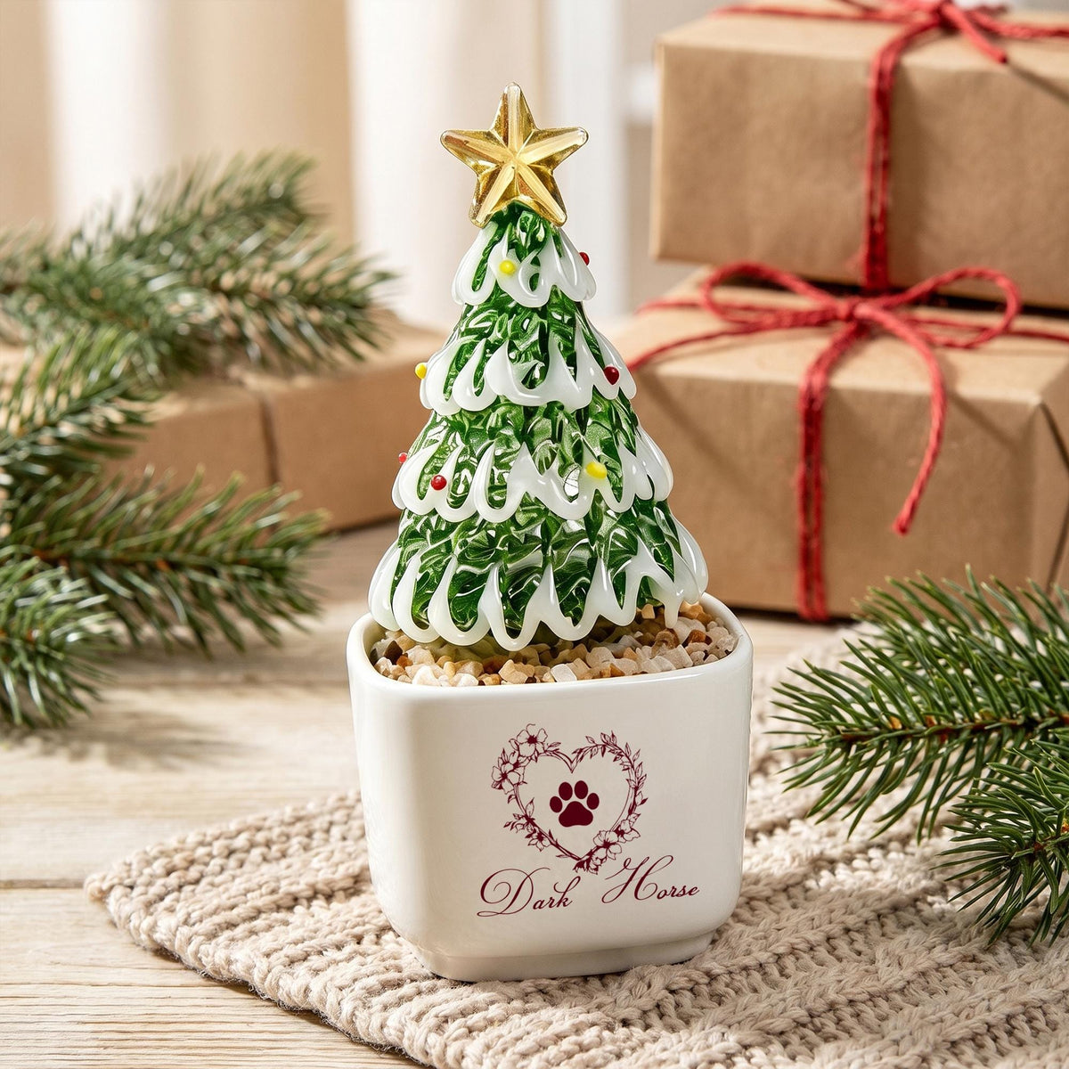 May include: A glass Christmas tree ornament with green and white swirls, topped with a gold star, sitting in a white square ceramic pot. The pot features a heart-shaped floral design with a paw print and the text Dark Horse printed in red.