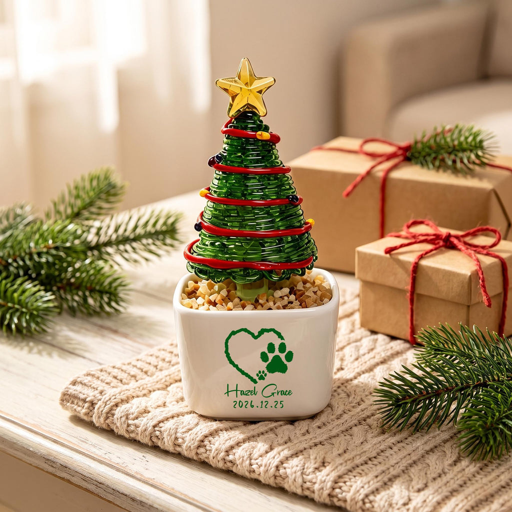 May include: A miniature green glass Christmas tree with a gold star topper, wrapped in red wire and small colored beads. The tree sits in a white square ceramic pot filled with pebbles, featuring a green heart, paw print, and the text Hazel Grace 2026.12.25.