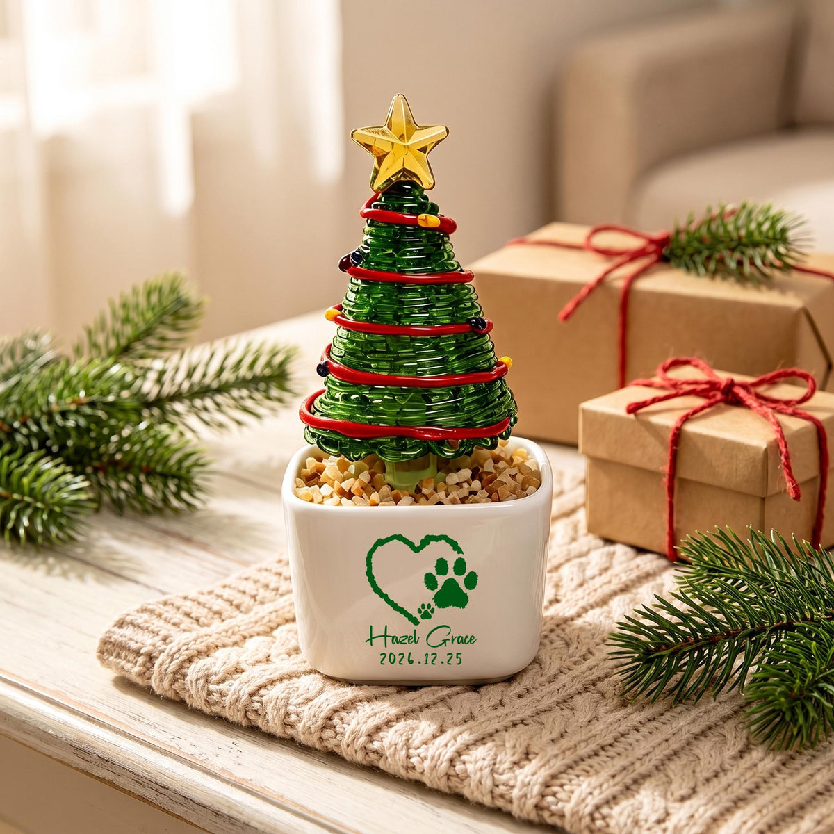 May include: A miniature green glass Christmas tree with a gold star topper, wrapped in red wire and small colored beads. The tree sits in a white square ceramic pot filled with pebbles, featuring a green heart, paw print, and the text Hazel Grace 2026.12.25.