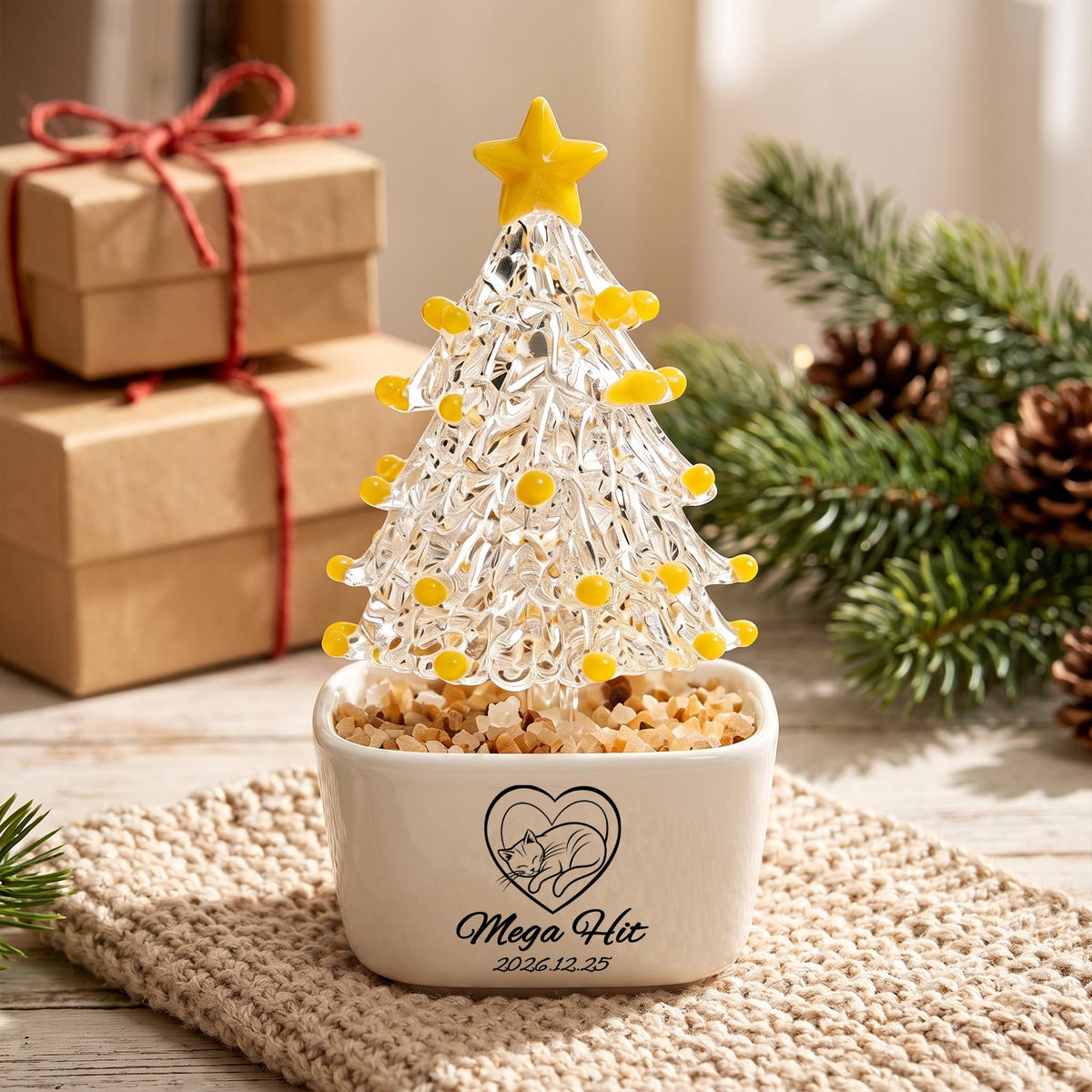 May include: A clear glass Christmas tree ornament with yellow glass accents and a yellow star topper, set in a white ceramic planter filled with small stones. The planter features a black line drawing of a cat inside a heart and the text Mega Hit 2026.12.25.