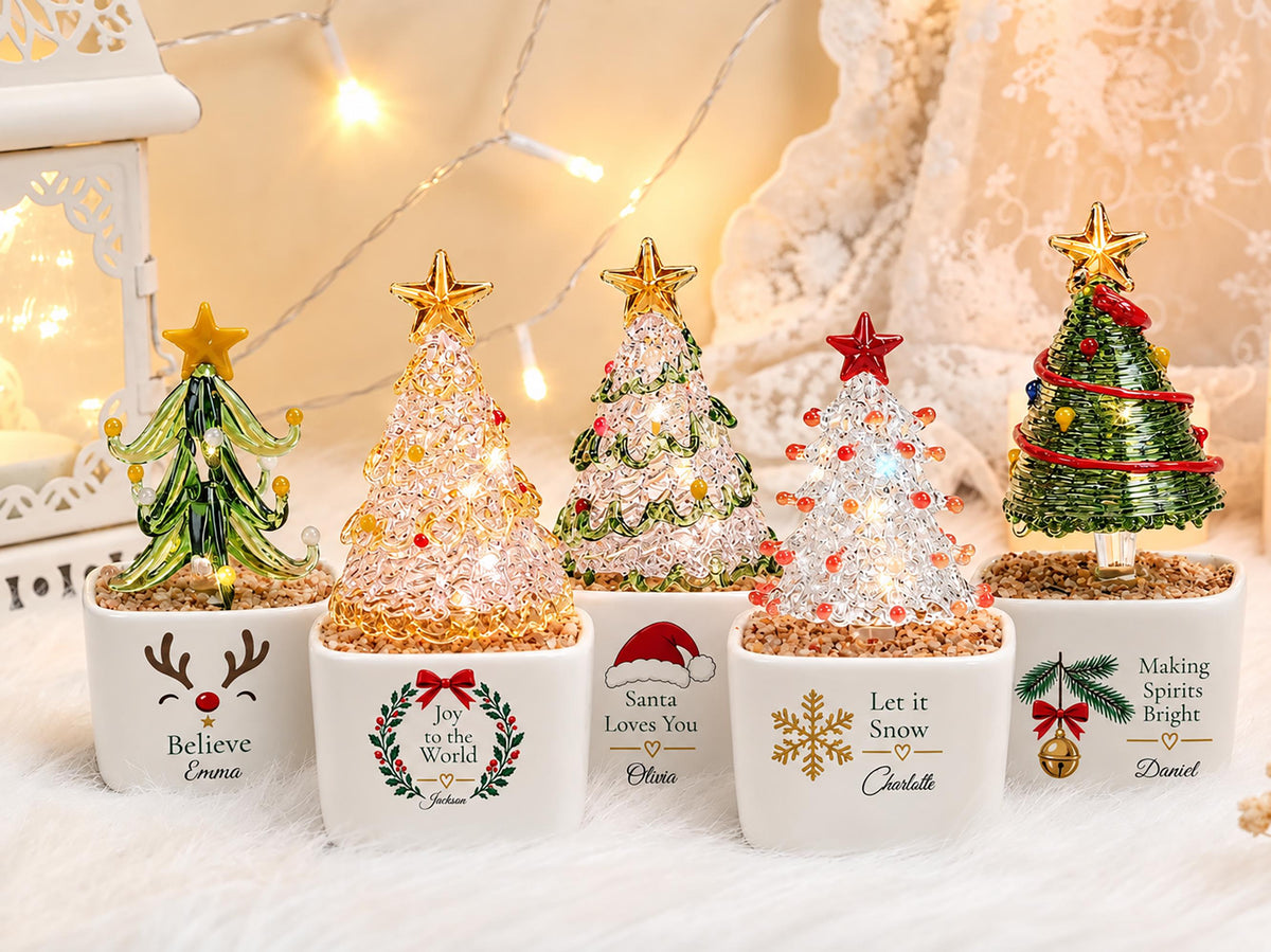 May include: Five miniature glass Christmas trees in white square ceramic pots. Each tree features unique decorations, including stars, lights, and festive graphics with names like Emma, Jackson, Olivia, Charlotte, and Daniel printed on the front.