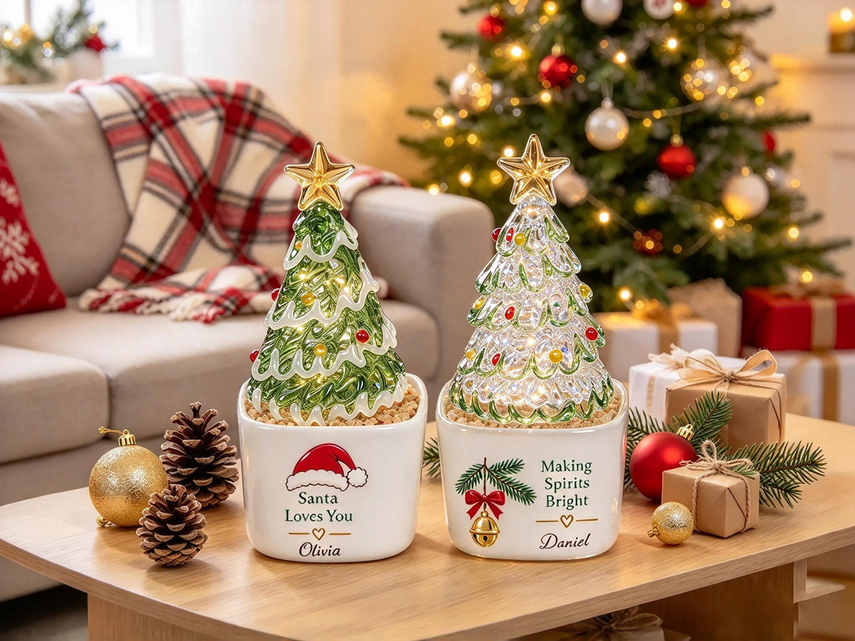 May include: Two ceramic planters containing glass Christmas trees with gold star toppers. The left planter features a Santa hat graphic and the text "Santa Loves You" with the name "Olivia". The right planter shows a bell graphic and "Making Spirits Bright" with the name "Daniel".