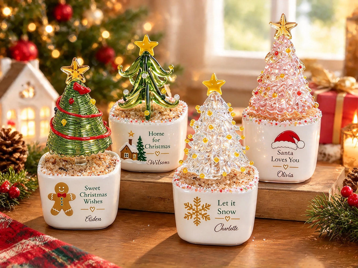 May include: Four glass Christmas tree figurines in white ceramic pots filled with sand. Each tree is topped with a yellow star and features unique decorations, including text like Sweet Christmas Wishes, Home for Christmas, Let it Snow, and Santa Loves You.