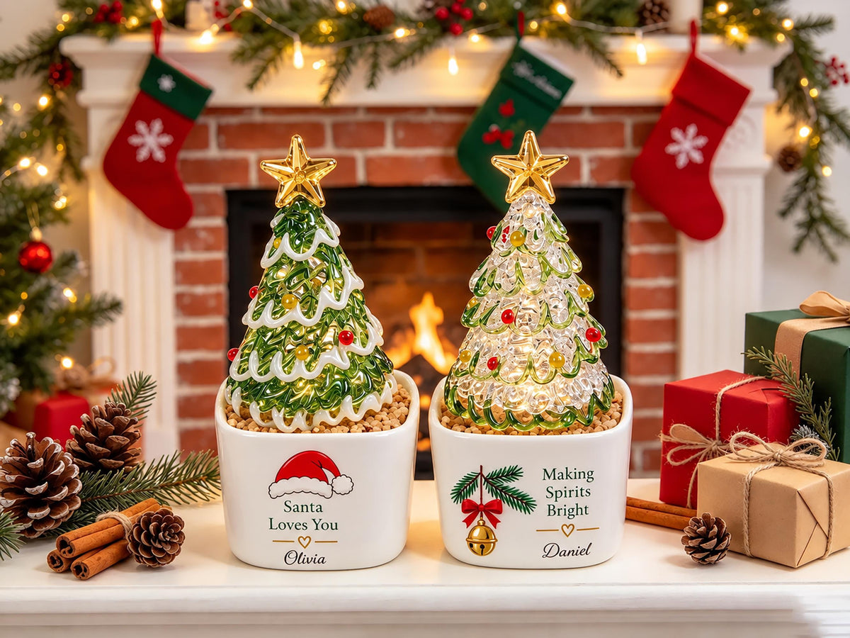 May include: Two white ceramic planters containing glass Christmas trees with gold stars. The left planter features a red Santa hat and the text "Santa Loves You" with the name "Olivia". The right planter shows a bell and "Making Spirits Bright" with "Daniel".