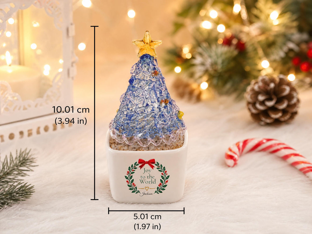 May include: A blue glass Christmas tree figurine with a yellow star topper, set in a white square ceramic planter. The planter features a wreath graphic with the text "Joy to the World" and measures 1.97 inches wide and 3.94 inches tall.