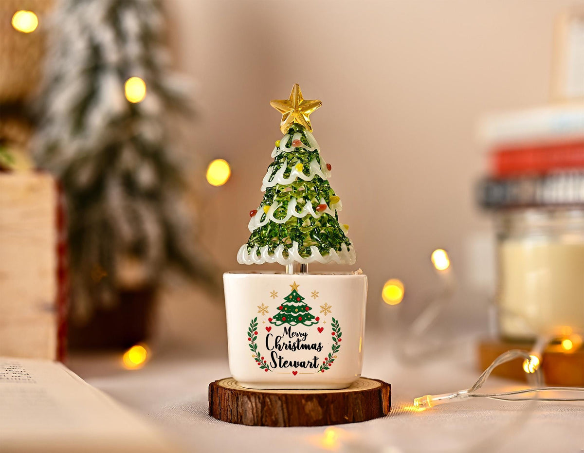May include: A decorative glass Christmas tree ornament with a gold star topper, featuring green and white glass branches, set in a white ceramic base printed with a green tree graphic, red hearts, and the text Merry Christmas Stewart.