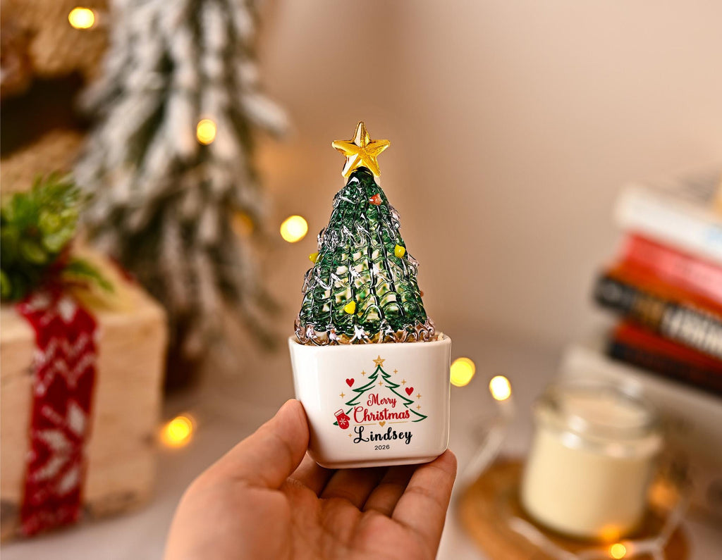 May include: A small, green, textured glass Christmas tree ornament with a gold star topper, set in a white ceramic pot. The pot features a printed green tree graphic, red hearts, and the text Merry Christmas, Lindsey, 2020 in black lettering.