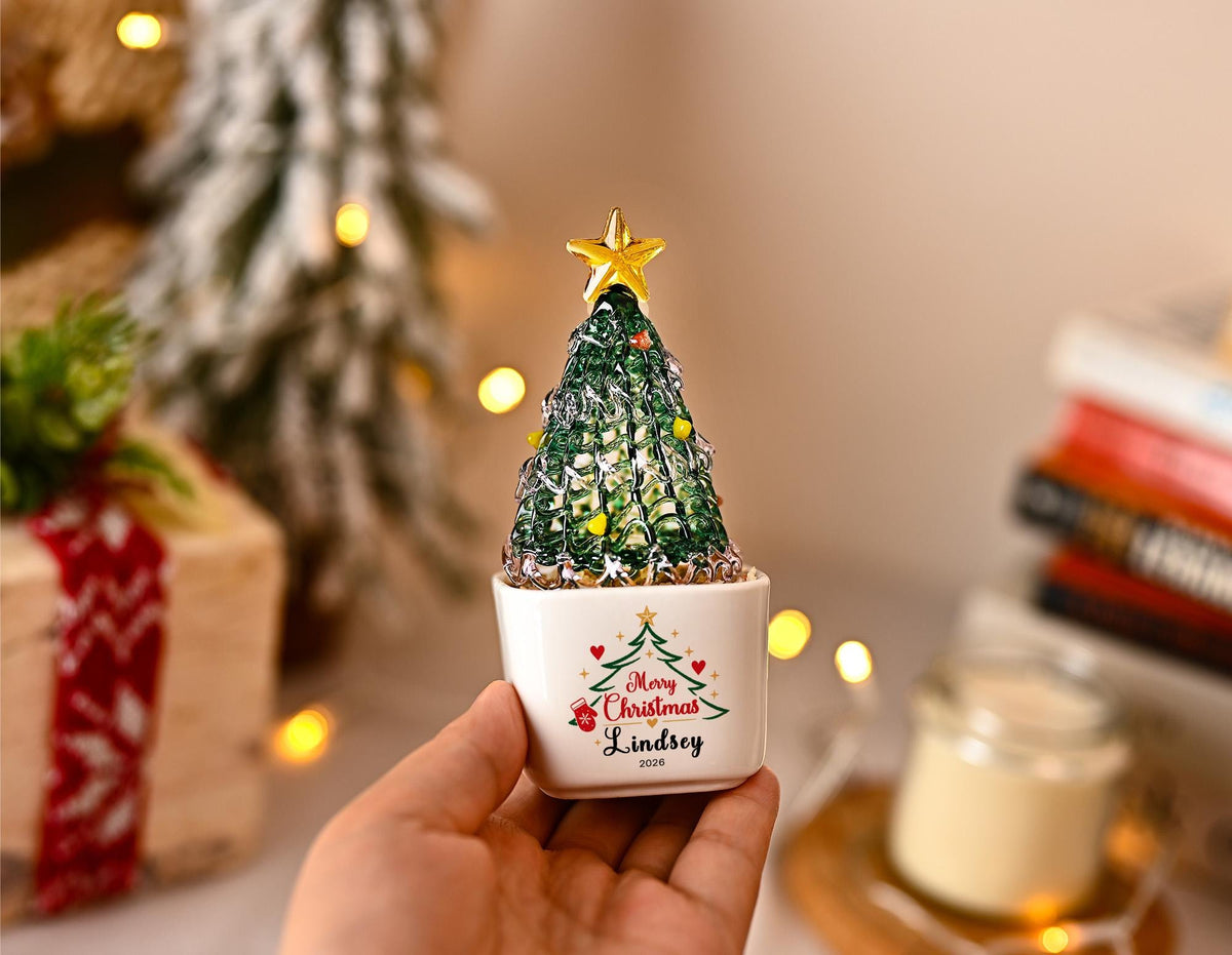 May include: A small, green, textured glass Christmas tree ornament with a gold star topper, set in a white ceramic pot. The pot features a printed green tree graphic, red hearts, and the text Merry Christmas, Lindsey, 2020 in black lettering.