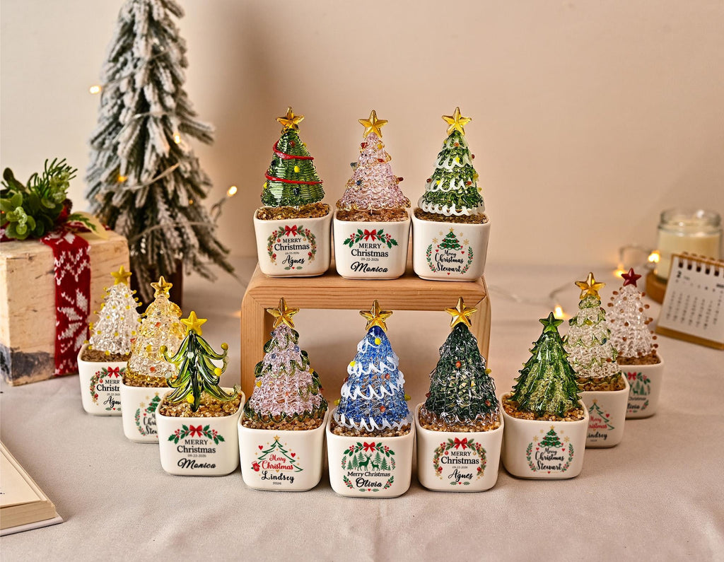 May include: A collection of small, decorative glass Christmas trees in white ceramic pots. Each tree features a gold star topper and sits on a bed of small stones. The pots are printed with a wreath design and the text Merry Christmas with names.