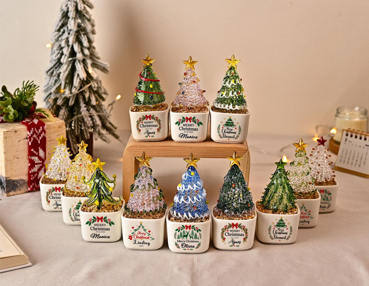 May include: A collection of small, decorative glass Christmas trees in white ceramic pots. Each tree features a gold star topper and sits on a bed of small stones. The pots are printed with a wreath design and the text Merry Christmas with names.