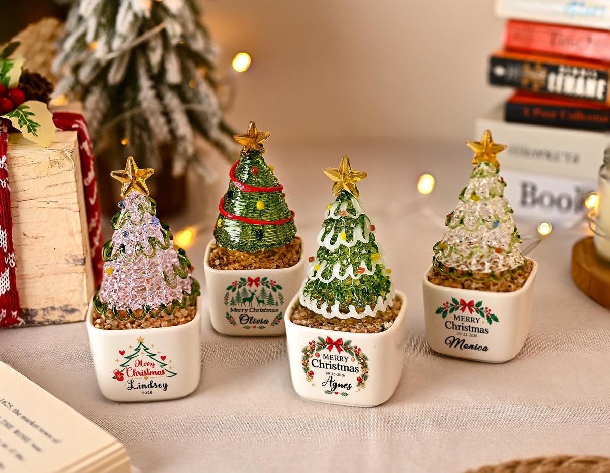 May include: Four small decorative Christmas tree figurines in white square ceramic pots. Each tree is crafted from textured glass with a gold star topper. The pots feature printed holiday graphics and names including Lindsey, Olivia, Agnes, and Monica.
