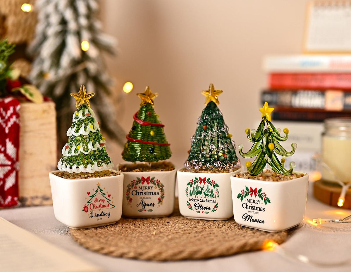 May include: Four miniature glass Christmas tree ornaments in white square ceramic pots. Each tree features a gold star topper and unique green glass designs. The pots are printed with Merry Christmas, names including Lindsey, Agnes, Olivia, and Monica, and festive holiday graphics.